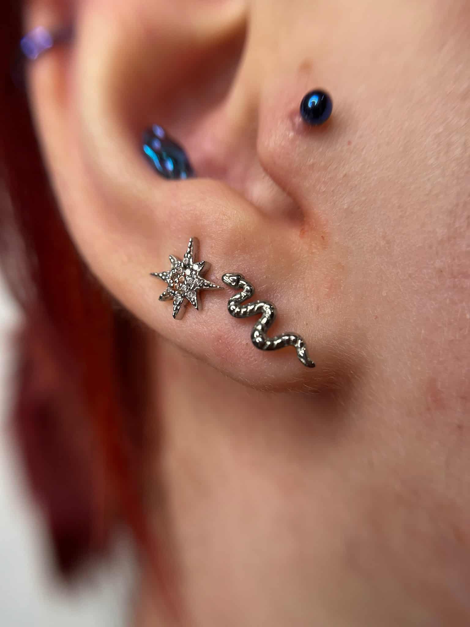 7sinstattoo.pl Zbliżenie na ucho z dwoma piercingami: jeden kolczyk w chrząstce ozdobiony niebieskim klejnotem i piercing płatka ucha ze srebrnym kolczykiem w kształcie węża wysadzanym małymi klejnotami, wszystko wykonane w studio tatuaży Poznań. Dodatkowo, kolejny mały piercing chrząstki z niebieskim klejnotem znajduje się bliżej głowy. Obraz jest oznaczony jako IMG_6664. Najlepsze studio tatuażu Seven Sins Tattoo