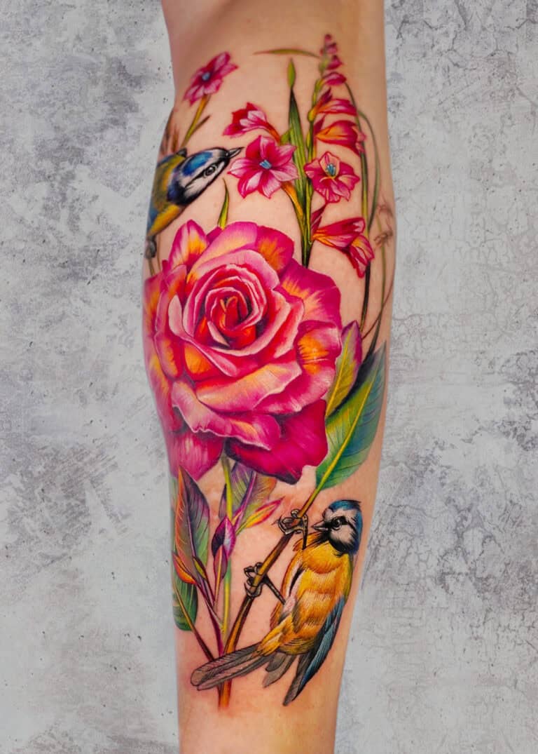 7sinstattoo.pl Żywy tatuaż na ramieniu osoby, wykonany w najlepszym studio tatuażu w Poznaniu, przedstawia dużą różową różę otoczoną mniejszymi różowymi kwiatami, zielonymi liśćmi i dwoma niebieskimi i żółtymi ptakami. Szczegółowy projekt ożywa dzięki realistycznej fakturze na neutralnym, teksturowanym szarym tle. Ta oszałamiająca grafika jest znana jako produkt x39. Najlepsze studio tatuażu Seven Sins Tattoo