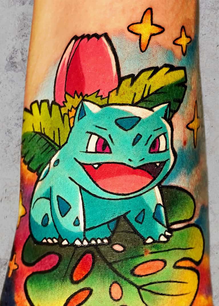 Kolorowy tatuaż na przedramieniu przedstawia ilustrację Bulbasaura, postaci Pokémon, z zielonymi liśćmi i różowym pączkiem na plecach. Tło tego tatuażu w kolorze obejmuje żywe kolory, gwiazdy i liściaste elementy.