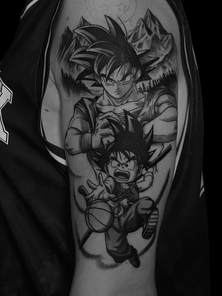 „tatuaz dragon ball songo” na górnym ramieniu przedstawia dwie postacie z kolczastymi włosami. Górna postać, z intensywnym wyrazem twarzy, stoi na tle górzystego krajobrazu. Poniżej, mniejsza postać wydaje się energiczna, trzymając piłkę i laskę — wykonana pieczołowicie w studiu tatuażu w Poznaniu.